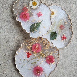 May include: Three white resin coasters with gold edges and embedded dried flowers. The coasters feature pink and white flowers, green leaves, and gold flakes.
