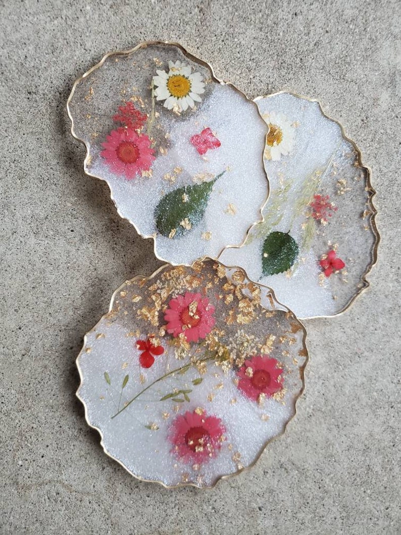 May include: Three white resin coasters with gold edges and embedded dried flowers. The coasters feature pink and white flowers, green leaves, and gold flakes.