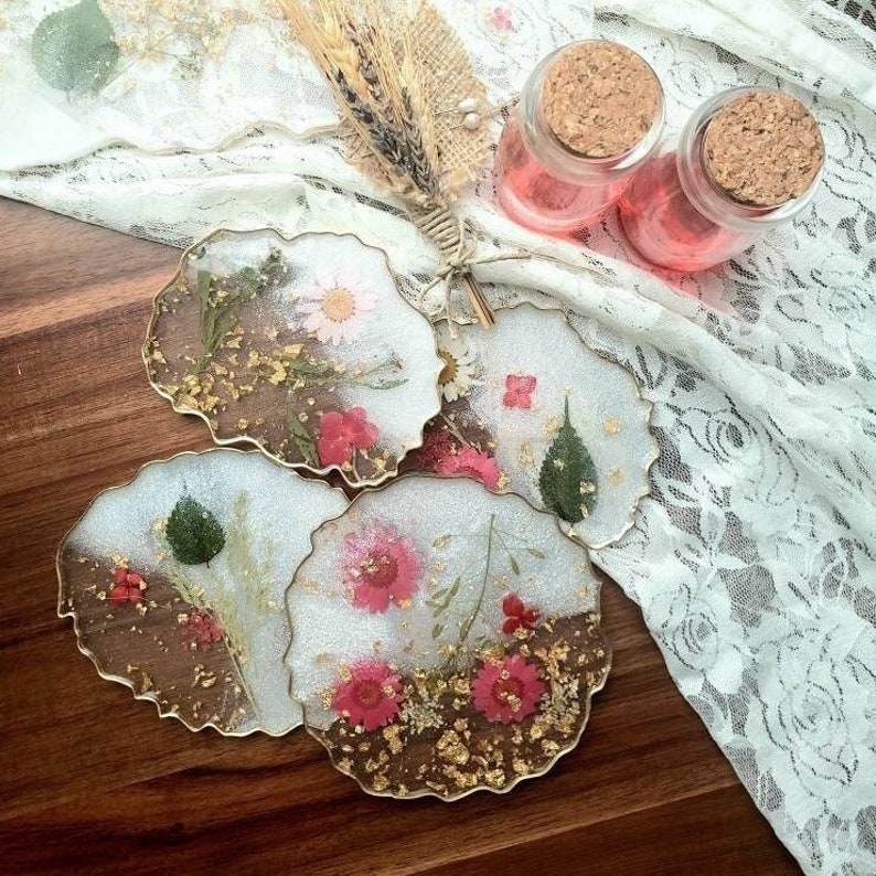 May include: Four round resin coasters with gold foil and pressed flowers. The coasters are white with a clear resin top and have a scalloped edge.