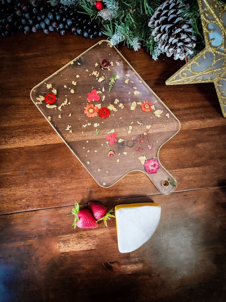 Op de afbeelding: Een transparant serveerbord met een gouden rand en gedroogde bloemen ingebed in de hars. Het bord heeft een handvat en ligt op een houten oppervlak. Er is een stuk witte kaas en aardbeien op het oppervlak.