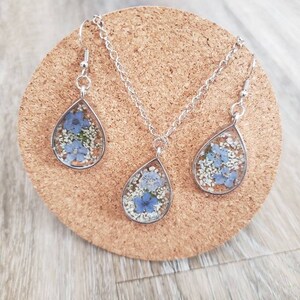 May include: A silver necklace and earrings set with teardrop-shaped pendants filled with pressed blue and white flowers.