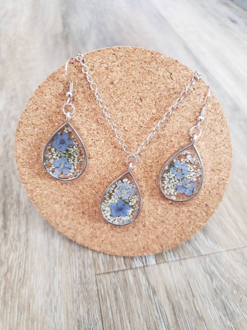 May include: A silver necklace and earrings set with teardrop-shaped pendants filled with pressed blue and white flowers.