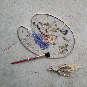 May include: A gold-rimmed, round, white resin tray with dried flowers and gold leaf embedded in the resin. A paintbrush is lying next to the tray. A small bundle of dried wheat is on the grey concrete surface.