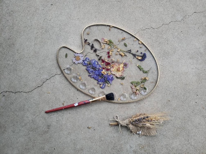 May include: A gold-rimmed, round, white resin tray with dried flowers and gold leaf embedded in the resin. A paintbrush is lying next to the tray. A small bundle of dried wheat is on the grey concrete surface.