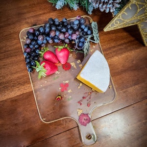 Op de afbeelding: Een transparant serveerbord met een handvat, versierd met gedroogde bloemen en bladgoud. Op het bord liggen een stuk kaas, druiven en aardbeien.