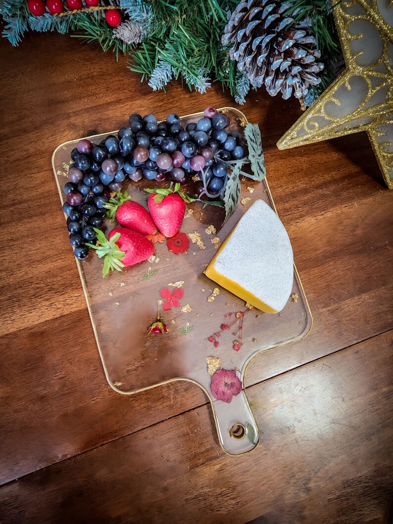 Op de afbeelding: Een transparant serveerbord met een handvat, versierd met gedroogde bloemen en bladgoud. Op het bord liggen een stuk kaas, druiven en aardbeien.