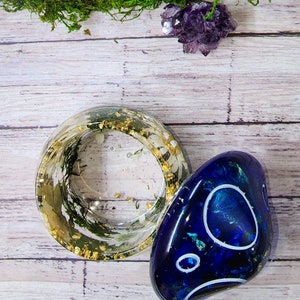 May include: A clear resin ring with gold flakes and green moss inside, next to a blue resin stone with white swirls.