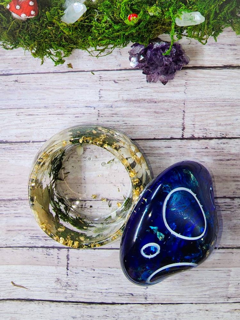May include: A clear resin ring with gold flakes and green moss inside, next to a blue resin stone with white swirls.