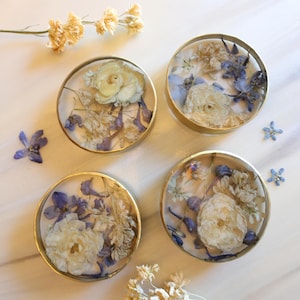 May include: Four round coasters with gold rims, each containing a unique arrangement of dried flowers in shades of white, cream, and purple. Delicate floral accents surround the coasters, creating a natural, elegant aesthetic.