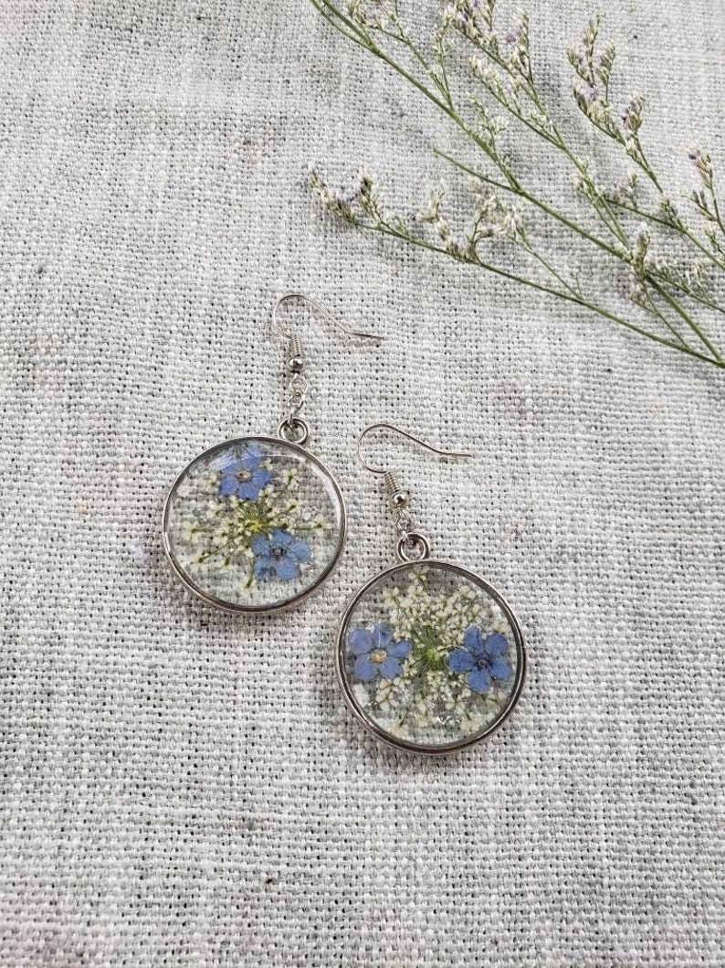 May include: A pair of silver earrings with round, clear resin pendants featuring pressed blue and white flowers.