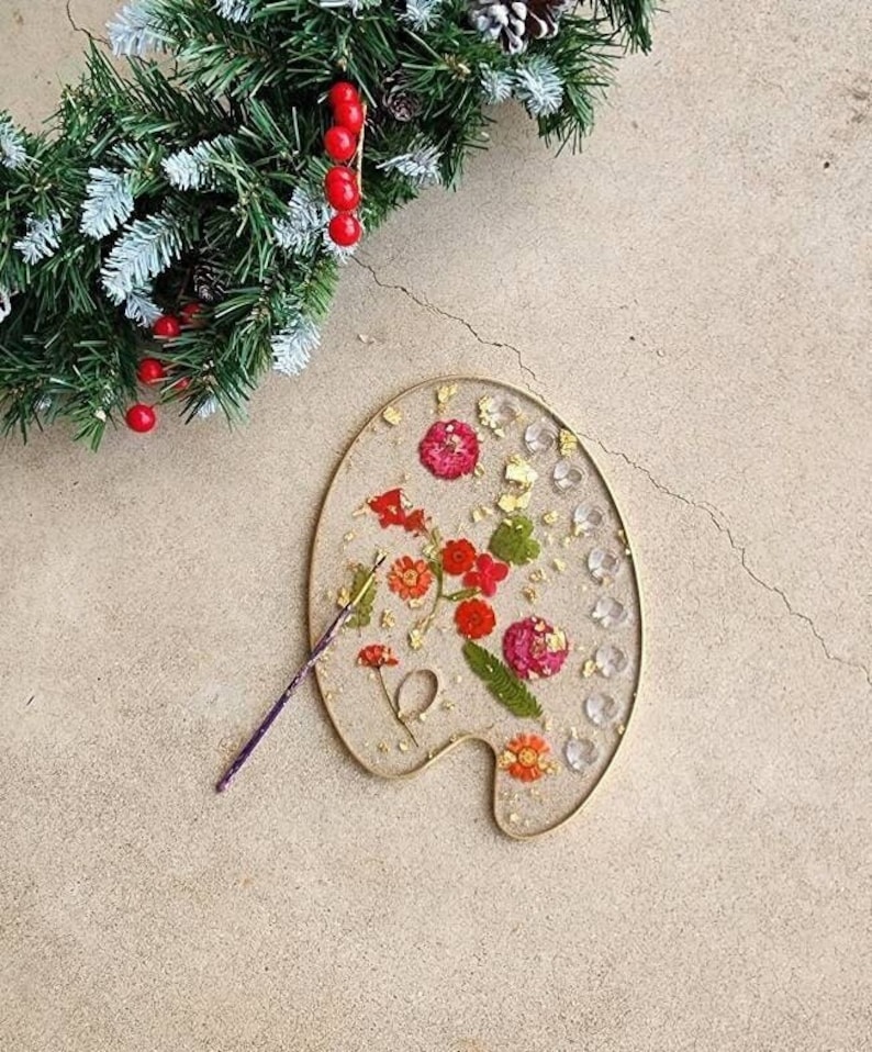 May include: A gold-rimmed artist's palette with a clear resin top. The resin contains pressed flowers and gold leaf. A paintbrush is lying next to the palette.