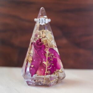 May include: A clear, geometric ring holder with dried flowers and gold flakes encased in resin. A silver ring with a large, clear centre stone sits atop the holder. The holder features deep pink rose petals.