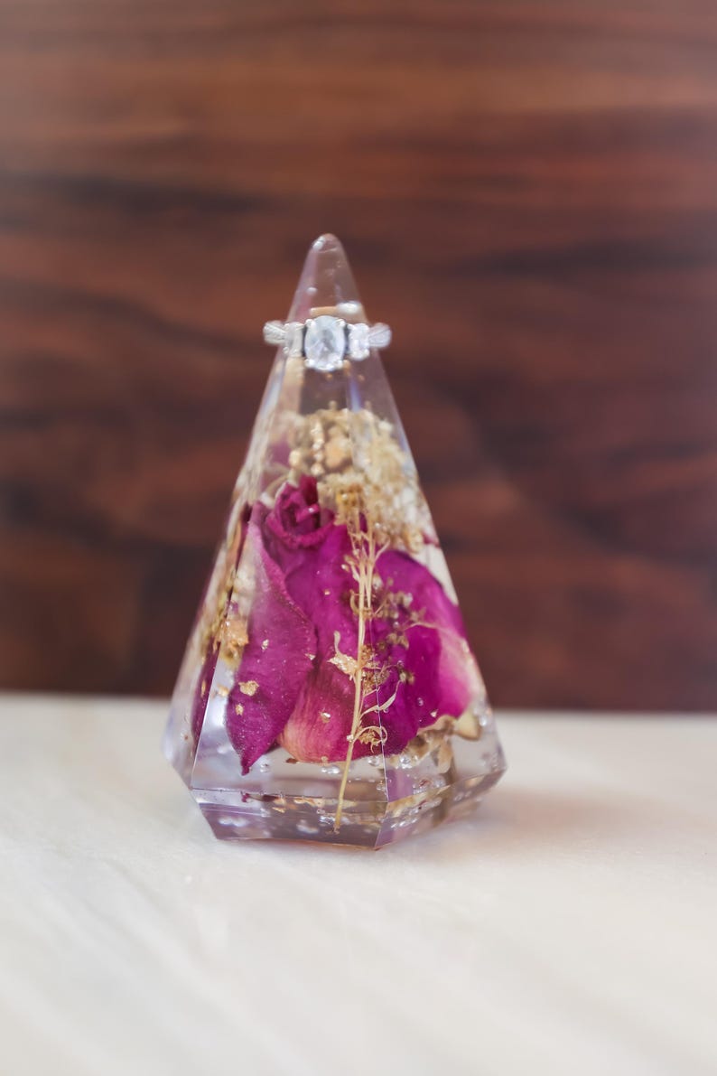 May include: A clear, geometric ring holder with dried flowers and gold flakes encased in resin. A silver ring with a large, clear centre stone sits atop the holder. The holder features deep pink rose petals.