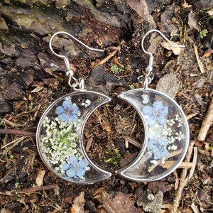 May include: A pair of silver crescent moon earrings with blue and white pressed flowers encased in clear resin.