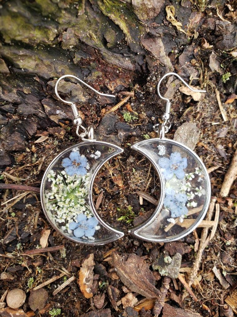 May include: A pair of silver crescent moon earrings with blue and white pressed flowers encased in clear resin.