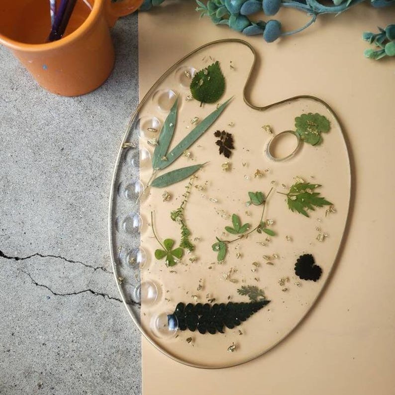 May include: A clear resin artist's palette with a gold rim, featuring pressed leaves and flowers, gold flakes, and a black heart shape.
