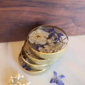 May include: A stack of four gold-colored coasters, each filled with dried flowers in shades of white, blue, and beige. The coasters are arranged on a light-colored surface, with a wooden background.