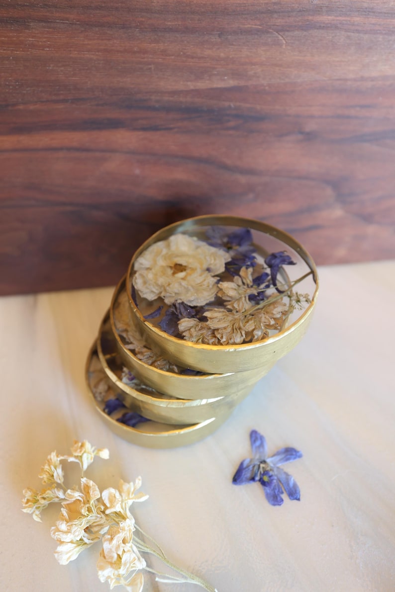May include: A stack of four gold-colored coasters, each filled with dried flowers in shades of white, blue, and beige. The coasters are arranged on a light-colored surface, with a wooden background.
