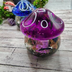 May include: Two resin mushroom lamps with a clear base filled with dried flowers and gold flakes. The mushroom on the left is blue with white accents and the mushroom on the right is purple with white accents.