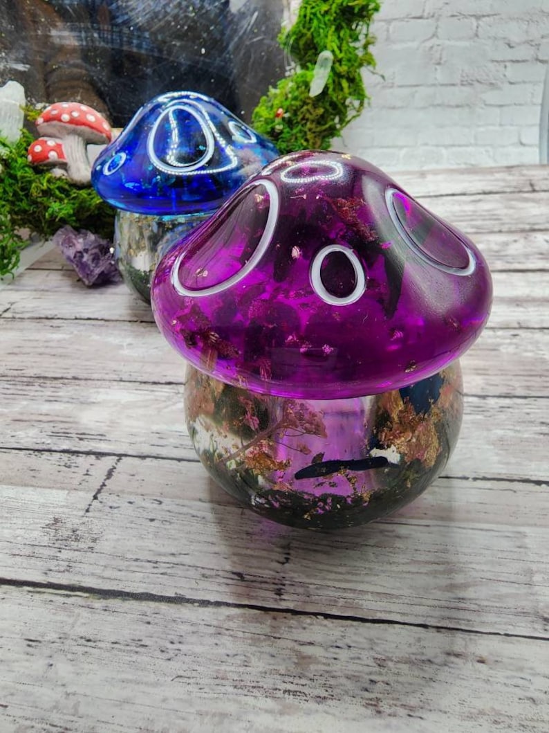 May include: Two resin mushroom lamps with a clear base filled with dried flowers and gold flakes. The mushroom on the left is blue with white accents and the mushroom on the right is purple with white accents.