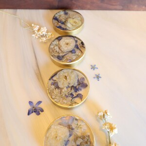 May include: Four round coasters with gold rims, each containing a unique arrangement of dried white and blue flowers. The coasters are arranged in a row on a white marble surface, with additional flowers scattered around.