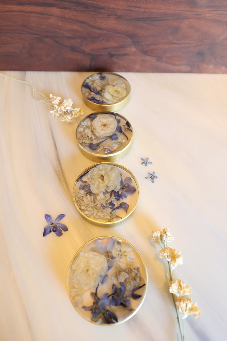 May include: Four round coasters with gold rims, each containing a unique arrangement of dried white and blue flowers. The coasters are arranged in a row on a white marble surface, with additional flowers scattered around.