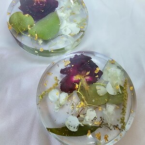 May include: Two clear resin coasters with embedded dried flowers and gold leaf flakes. Each coaster features a deep burgundy flower, white blossoms, and green leaves. The coasters are round and sit on a white surface.