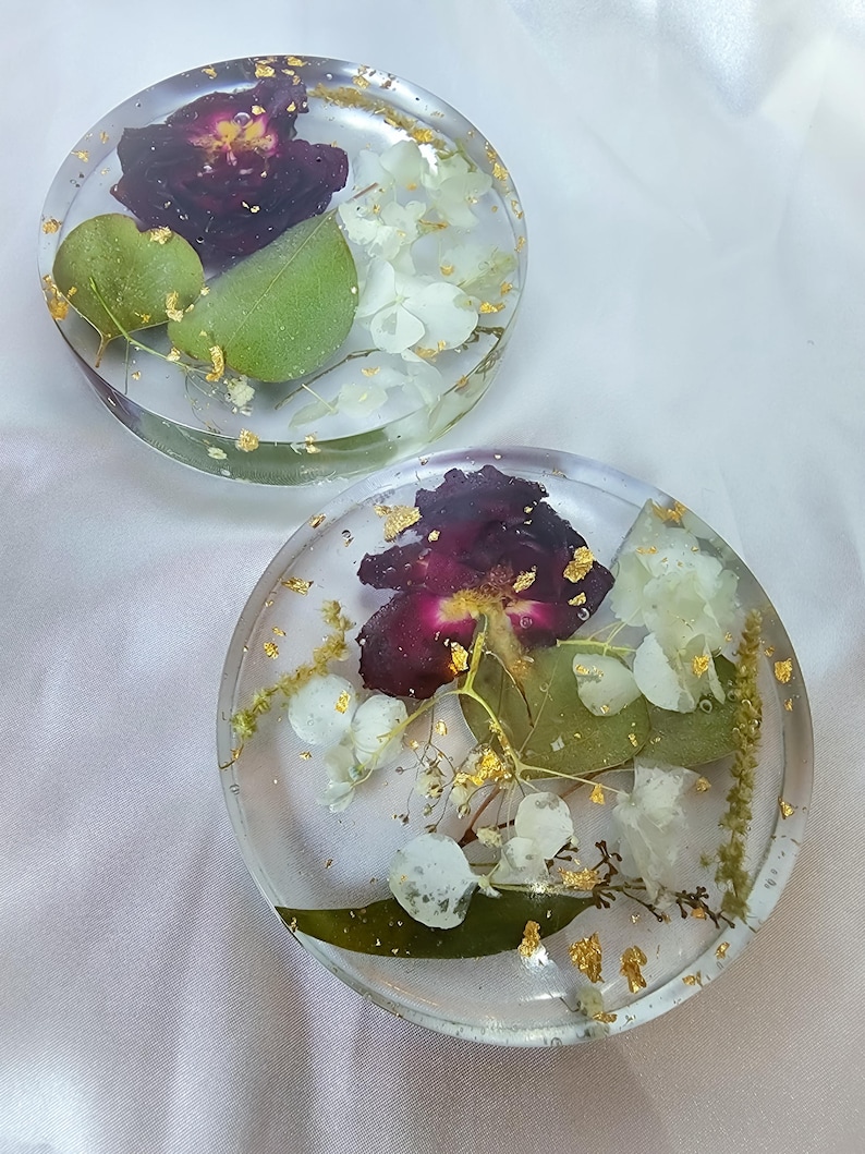 May include: Two clear resin coasters with embedded dried flowers and gold leaf flakes. Each coaster features a deep burgundy flower, white blossoms, and green leaves. The coasters are round and sit on a white surface.