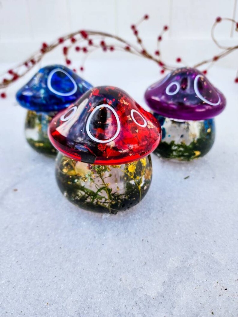 May include: Three resin mushroom ornaments with different colored tops. The mushrooms are filled with dried flowers and glitter. The tops are blue, red, and purple.