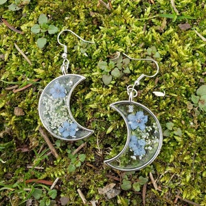 May include: A pair of silver crescent moon earrings with delicate blue and white pressed flowers encased in clear resin.