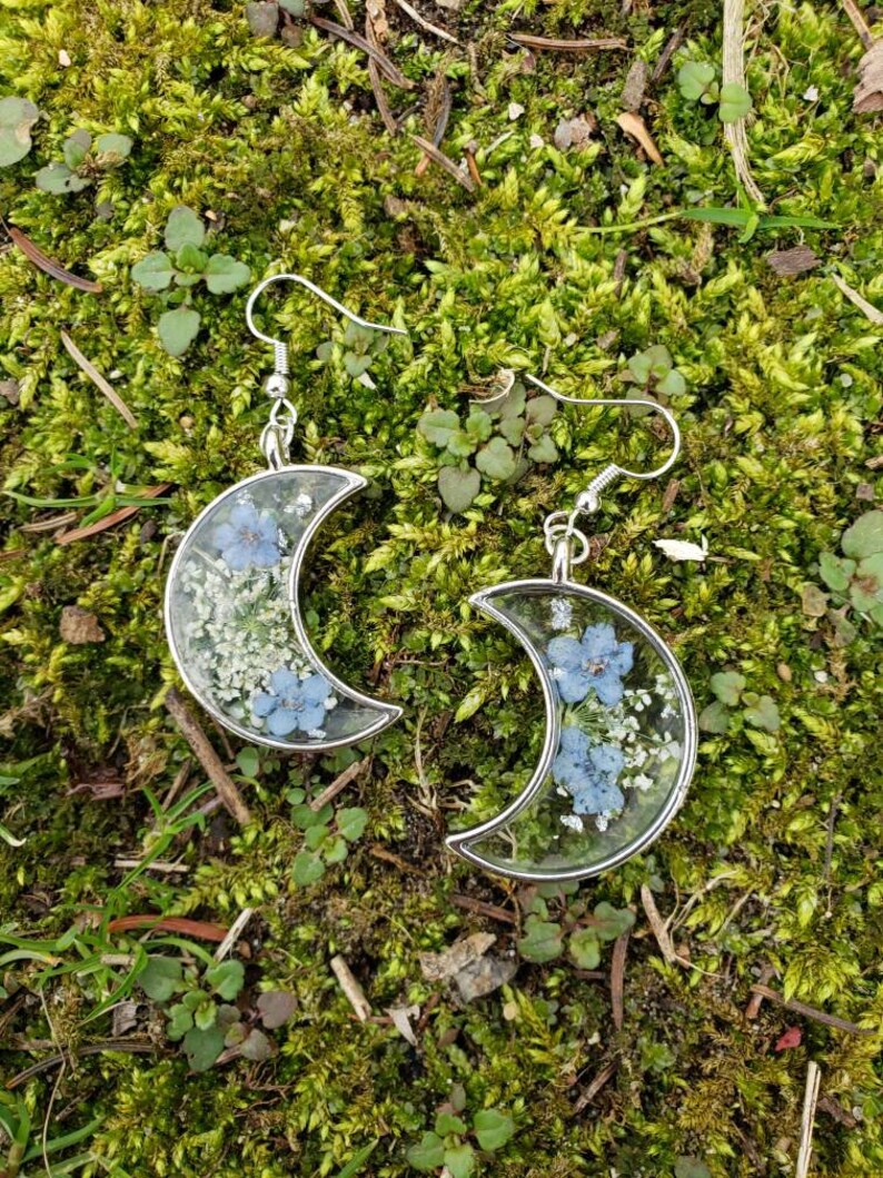 May include: A pair of silver crescent moon earrings with delicate blue and white pressed flowers encased in clear resin.