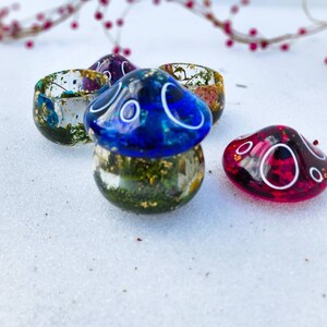 May include: Three small resin mushroom-shaped containers with gold flecks and dried flowers inside. The tops of the mushrooms are blue, pink, and red.