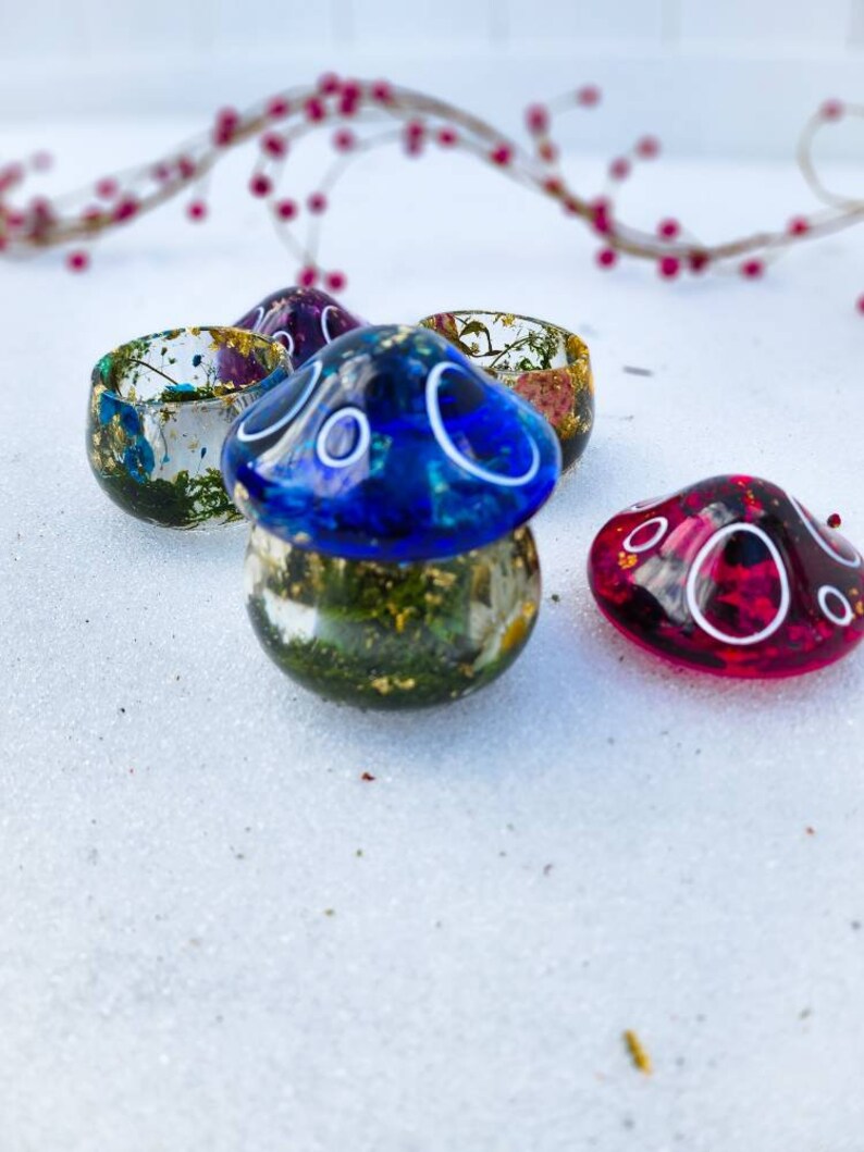 May include: Three small resin mushroom-shaped containers with gold flecks and dried flowers inside. The tops of the mushrooms are blue, pink, and red.