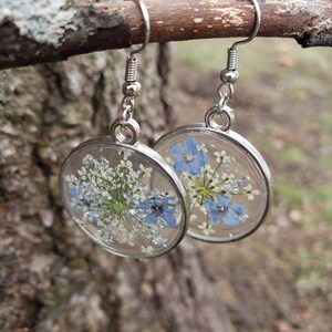 May include: A pair of silver dangle earrings with clear resin and pressed flowers. The flowers are blue and white and have a sparkly finish.