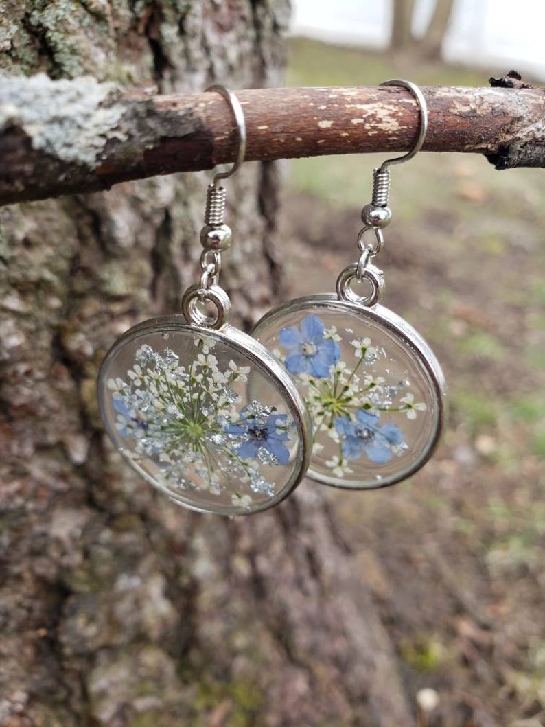 May include: A pair of silver dangle earrings with clear resin and pressed flowers. The flowers are blue and white and have a sparkly finish.