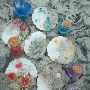 May include: Eight round, white resin coasters with gold trim and pressed flowers and gold leaf. The coasters are arranged in a circle on a gray textured surface. There are also six small glass bottles with cork stoppers.