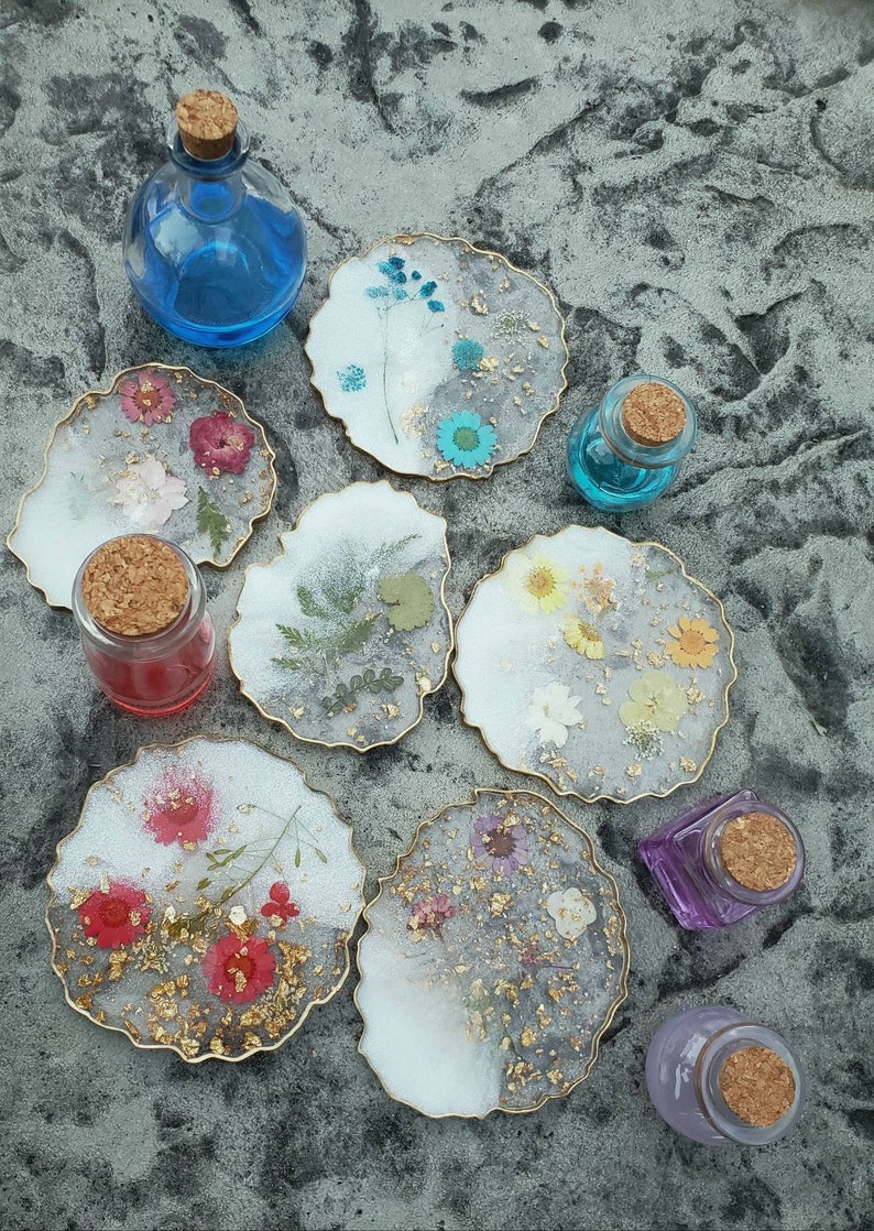 May include: Eight round, white resin coasters with gold trim and pressed flowers and gold leaf. The coasters are arranged in a circle on a gray textured surface. There are also six small glass bottles with cork stoppers.