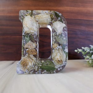May include: A clear resin letter D with dried white roses and greenery inside. The letter is sitting on a white surface.