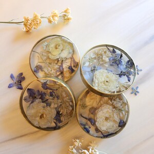 May include: Four round coasters with dried flowers encased in clear resin. The coasters have gold-colored metal rims and feature white and blue floral arrangements. A few loose dried flowers are scattered around the coasters.
