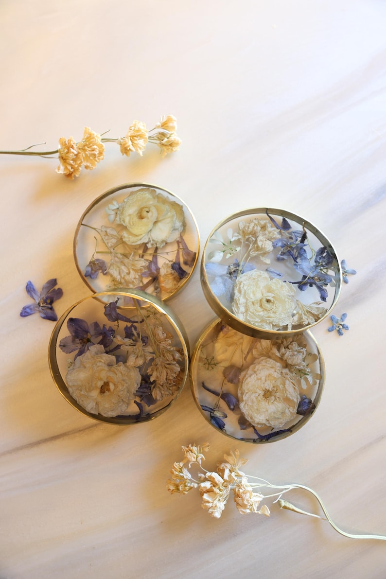 May include: Four round coasters with dried flowers encased in clear resin. The coasters have gold-colored metal rims and feature white and blue floral arrangements. A few loose dried flowers are scattered around the coasters.