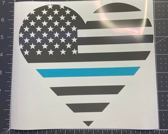 May include: A heart-shaped vinyl decal featuring a black and white American flag pattern with a horizontal thin blue line across the center. The decal is positioned on a cutting mat with a visible ruler showing a width of approximately 7.5 inches.
