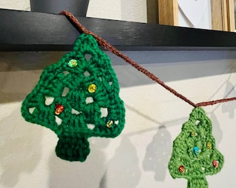 Könnte beinhalten: Eine grüne gehäkelte Weihnachtsbaumgirlande mit glitzernden Verzierungen. Die Girlande ist auf braunem Garn aufgefädelt und hängt an einem schwarzen Regal.