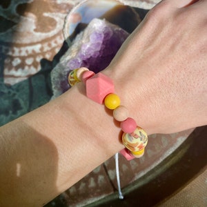 May include: A pink and yellow beaded bracelet with a floral pattern. The bracelet is made of silicone and has a square-shaped bead in the centre.