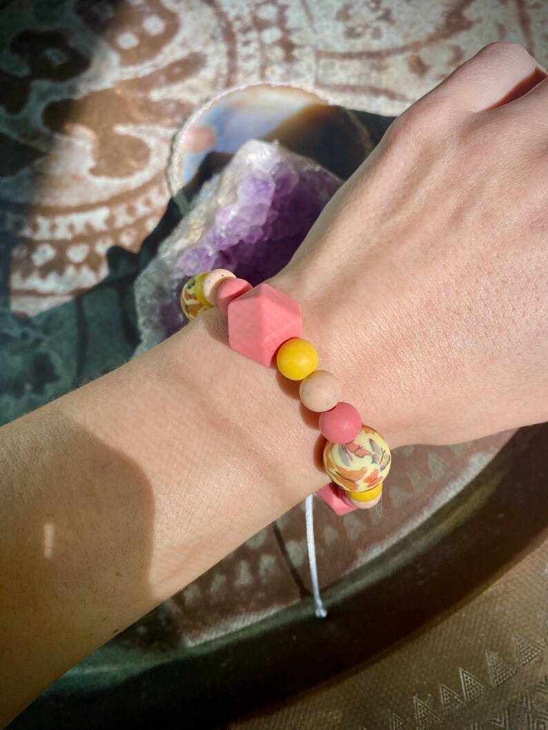 May include: A pink and yellow beaded bracelet with a floral pattern. The bracelet is made of silicone and has a square-shaped bead in the centre.