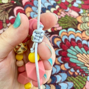 May include: A close-up of a light grey cord tied in a knot around a pink and yellow silicone bead bracelet. The bracelet has a variety of shapes and sizes of beads.