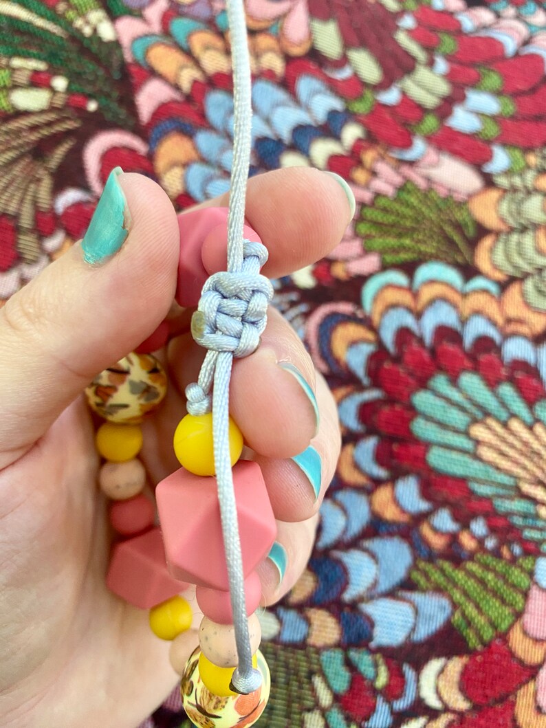 May include: A close-up of a light grey cord tied in a knot around a pink and yellow silicone bead bracelet. The bracelet has a variety of shapes and sizes of beads.
