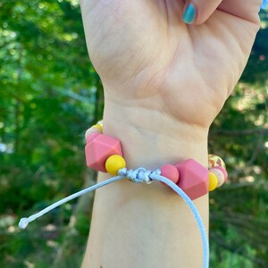 May include: A pink and yellow silicone bracelet with a knotted cord fastening. The bracelet is made of chunky beads with different shapes and textures.
