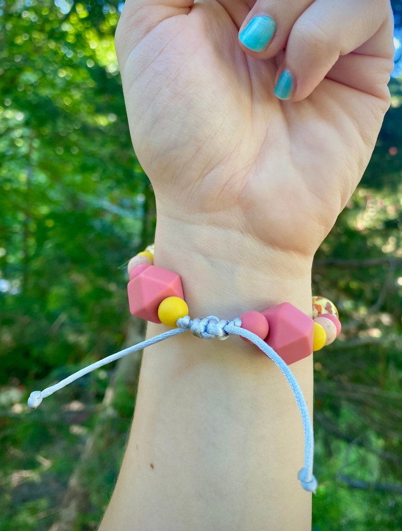 May include: A pink and yellow silicone bracelet with a knotted cord fastening. The bracelet is made of chunky beads with different shapes and textures.