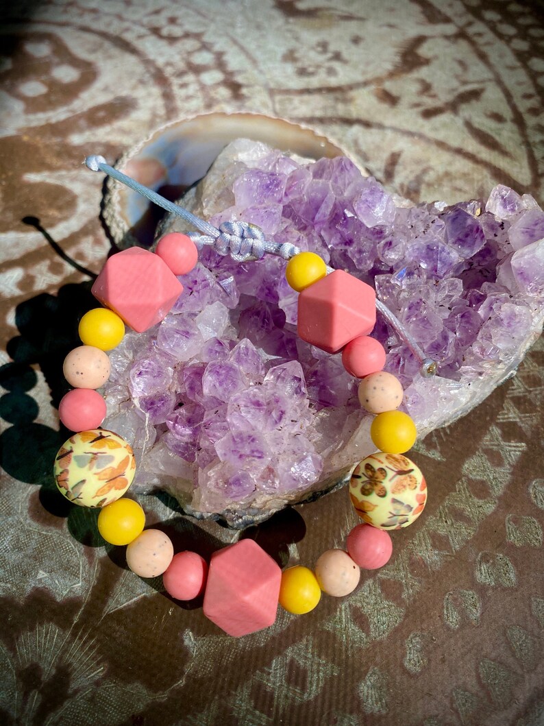 May include: A pink and yellow silicone bracelet with a floral pattern. The bracelet is made of multiple beads, including some with a geometric shape.