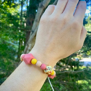 May include: A pink and yellow silicone bracelet with a floral pattern bead. The bracelet is on a person's wrist.